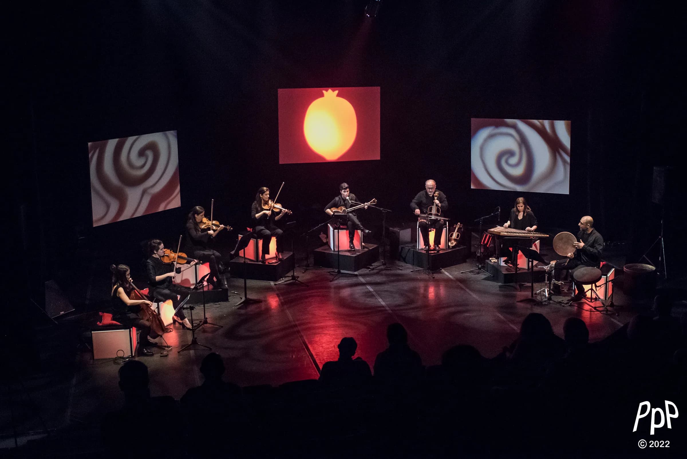 The ensemble seated in a wide arc on a darkened stage, a pomegranate projected on the screen behind them.