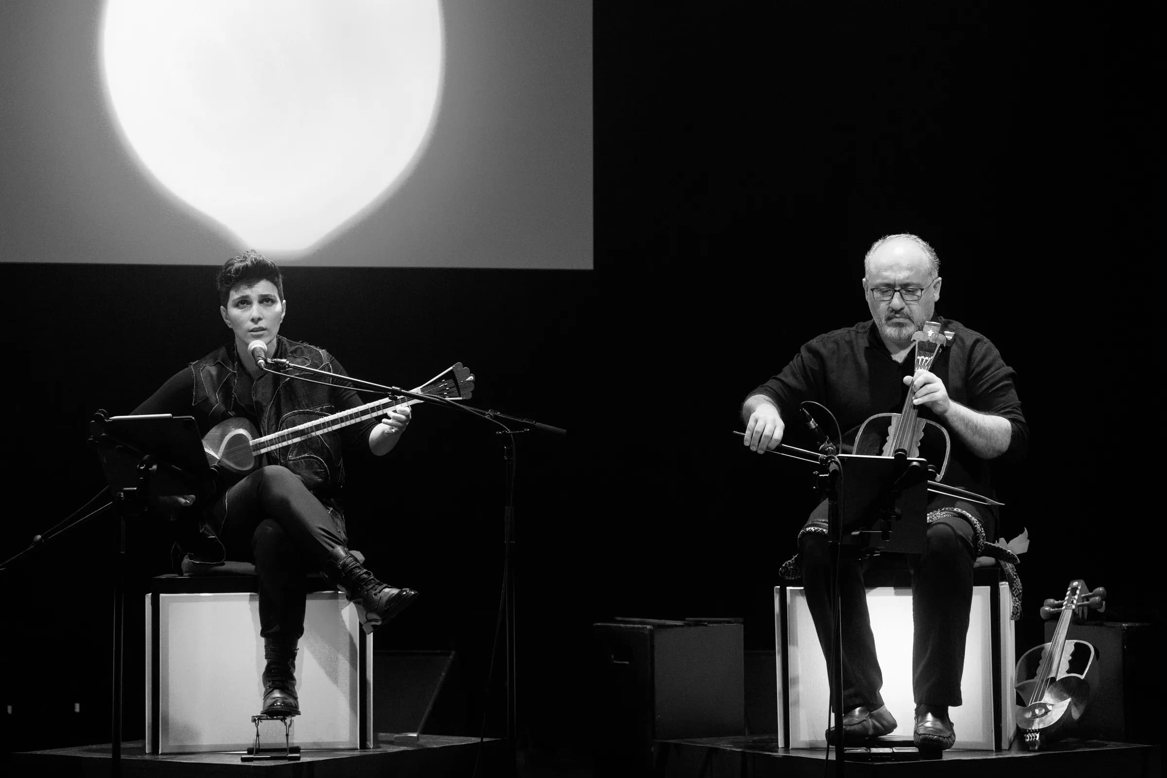 En noir et blanc : la chanteuse au târ et un joueur de gheychak, assis côte à côte sous un disque de lumière pâle.