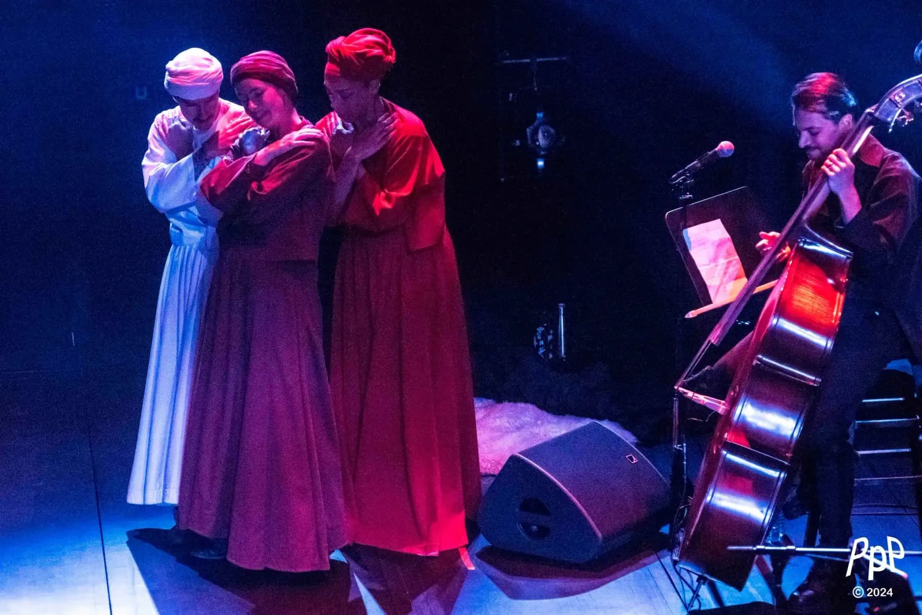 At the edge of the stage, three dancers in long robes and turbans held in one embrace; beside them, the double bass is played.