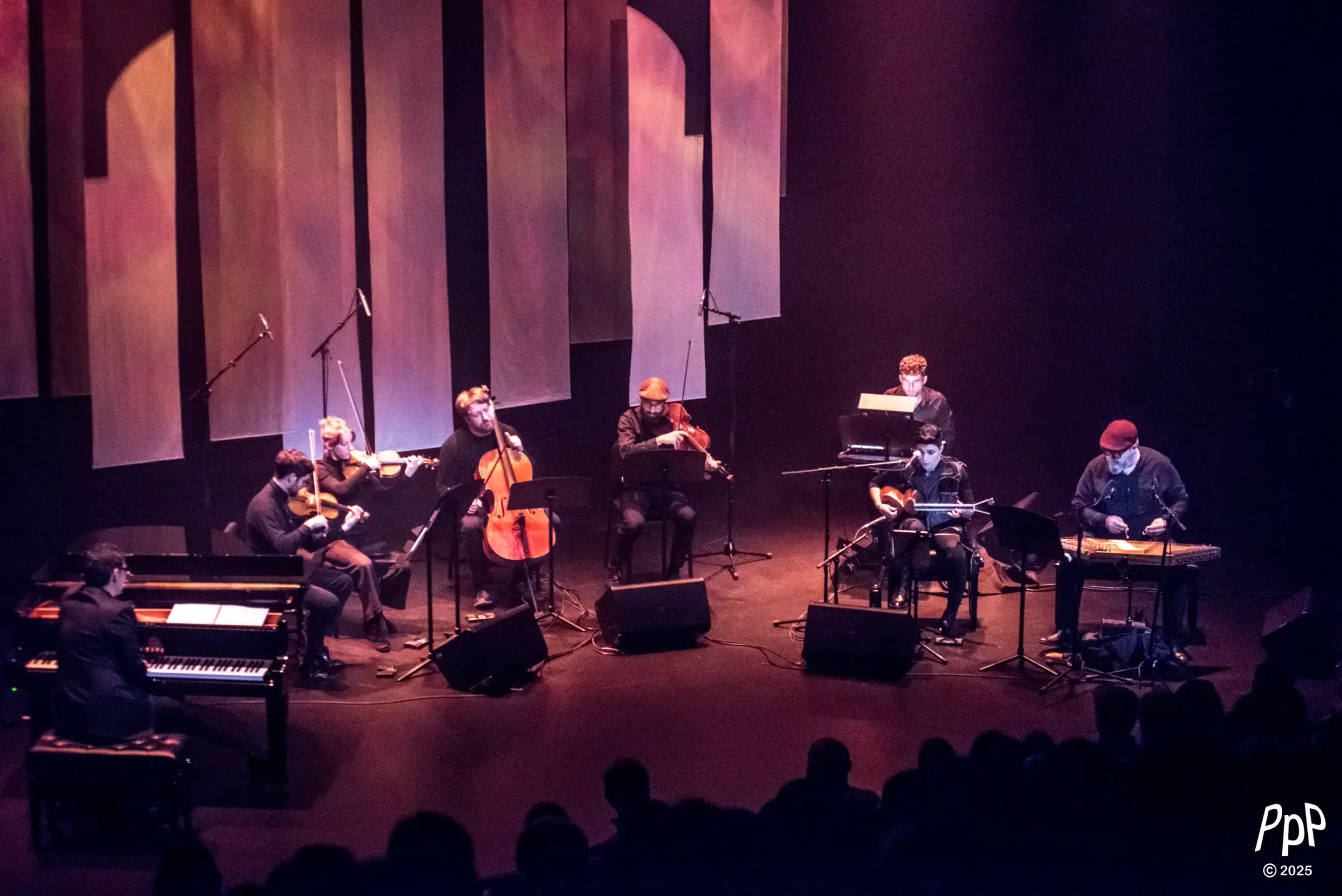The full ensemble on stage — piano, string quartet, synthesizer, târ and santur — beneath tall hanging panels lit in warm light.